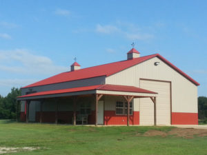 A large red and white barn stands on a grassy field under a clear blue sky. The structure features a high, red metal roof with two cupolas on top and a wide, vertical sliding door on one side. A covered porch area extends from the front.