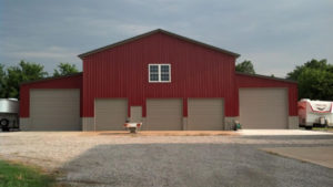 A large equipment storage pole barn with four closed garage doors and a single window at the center. The building is surrounded by gravel and pavement, with trees and a trailer visible in the background.