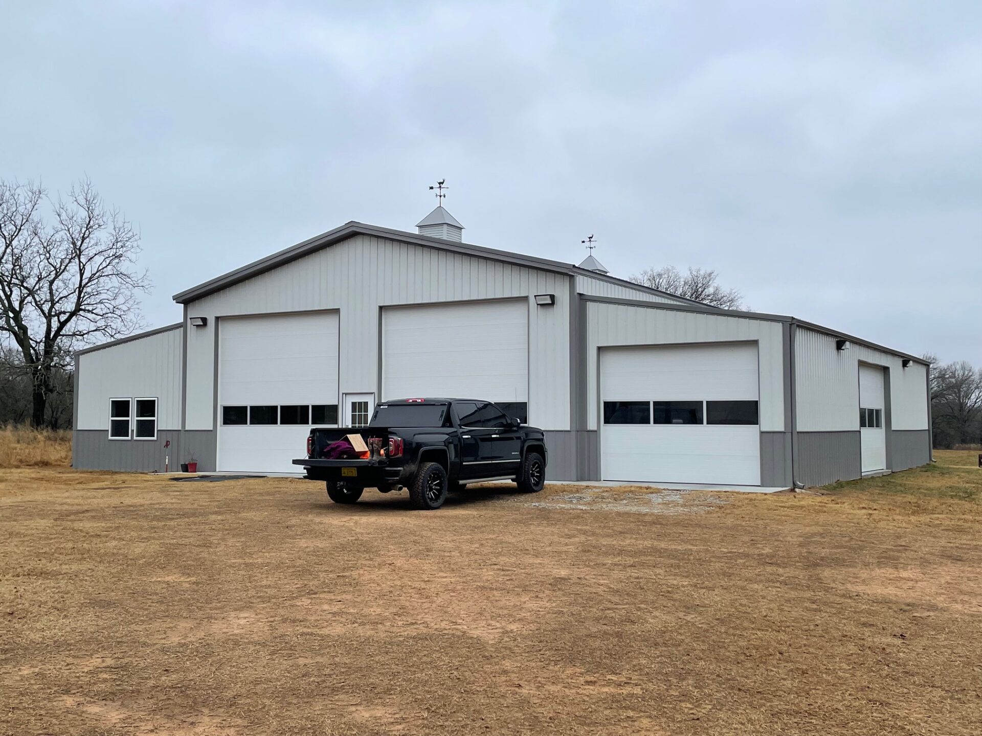 A large gray and white metal building with three garage doors and two small cupolas on the roof. A black pickup truck is parked in front of the building on a dirt lot. The surrounding area is grassy with trees in the background under an overcast sky.