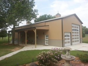 A large beige and brown barn with a white garage door and a small porch area on the left side. The porch has a seating area with chairs and a table. There is a manicured lawn with flowers and a birdbath in the foreground. Trees and a cloudy sky are in the background.