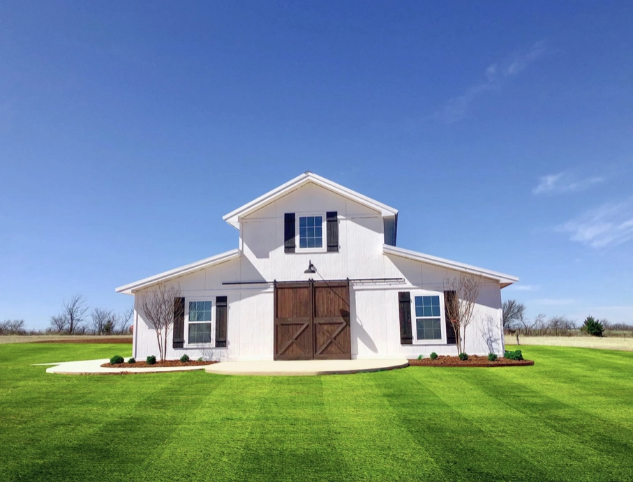 White barn with dark wood doors and windows, set in a large, neatly mowed green field. Clear blue sky above, with a few trees in the distance. Symmetrical design with two small trees and shrubs lining the entrance path.