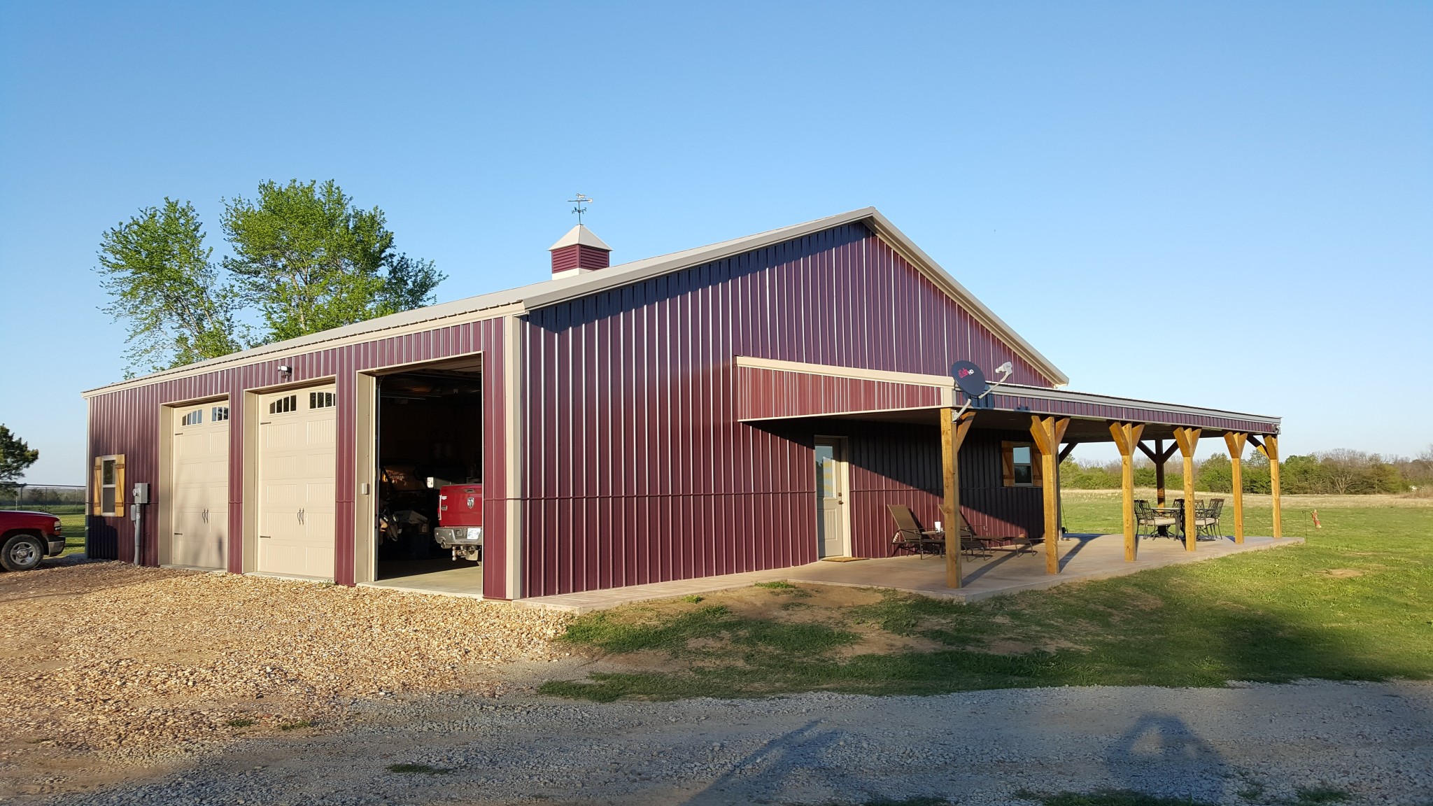 A large maroon metal post frame<br />
barn with beige garage doors, a small porch with wooden posts, and outdoor chairs sits on a gravel driveway in a grassy rural area under a clear blue sky.