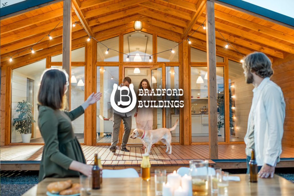 Two couples and a dog gather on the porch of a modern wooden house with string lights. Food and drinks are on the outdoor table. The Bradford Buildings logo is overlaid on the image.