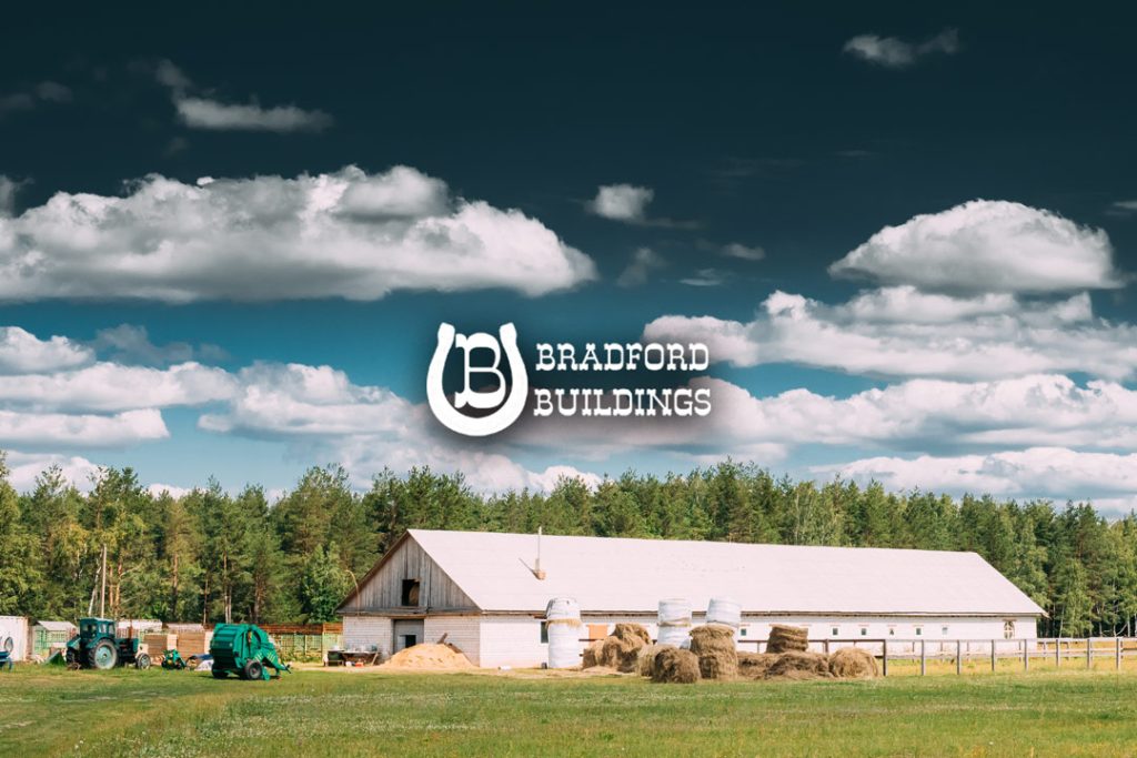 A large white barn sits on green grass with hay bales, farm equipment, and a forest in the background. The Bradford Buildings logo and name are centered in the sky above the barn.