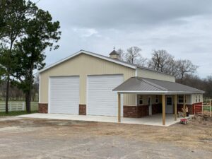 A large beige metal building with two tall garage doors, a covered porch, and a concrete driveway, set in a rural area with trees and a cloudy sky.